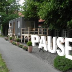 An einem begrünten Haus mit Tiny House ist das Wort PAUSE digital eingearbeitet.