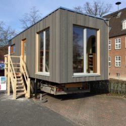 Ein Tiny House steht auf einen LKW-Anhänger vor einem Schulgebäude.