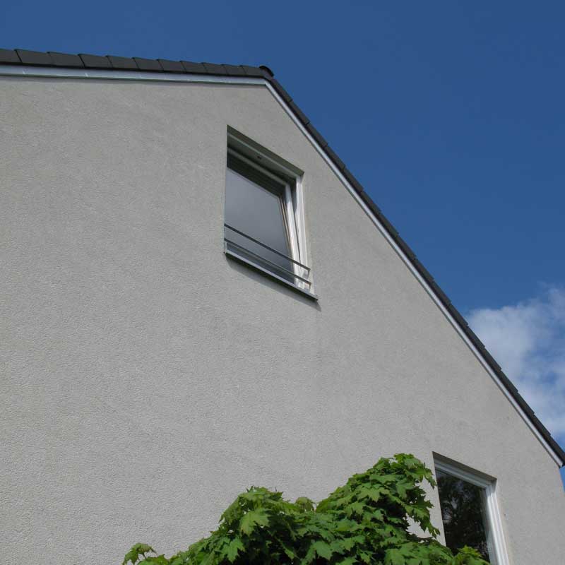 Untersicht auf den seitlichen Dachspitz mit einem hohen Fenster auf einer grauen Fassade.