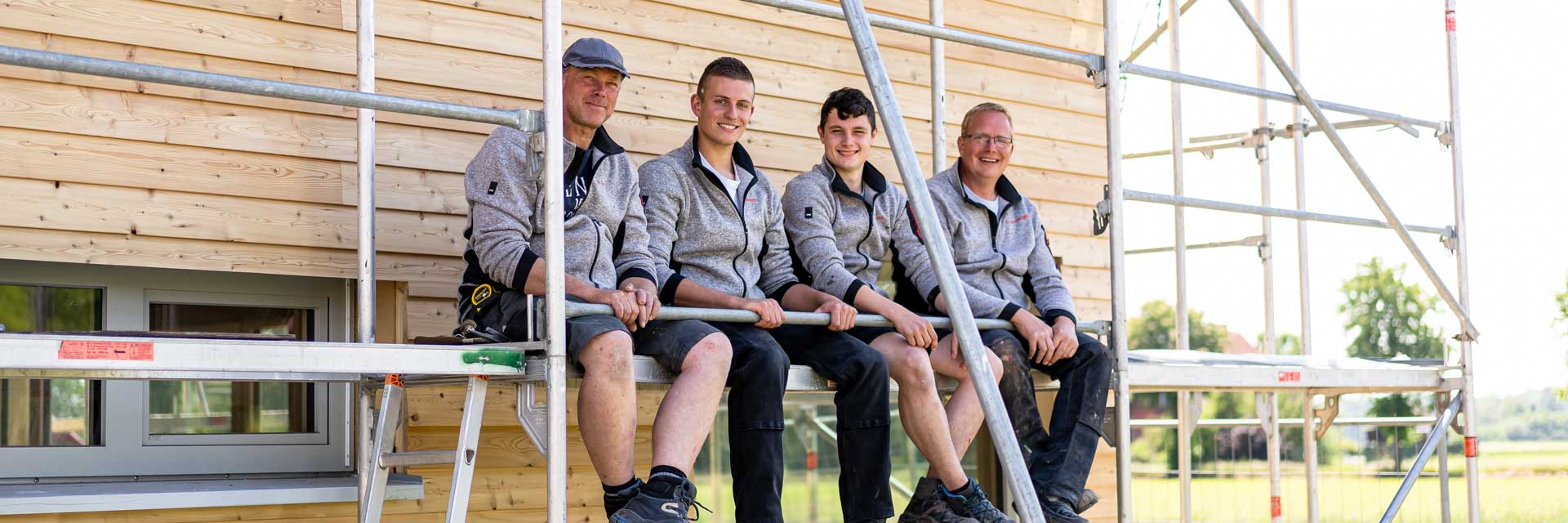 Vier Mitarbeiter sitzen lachend nebeneinander auf einem Baugerüst.